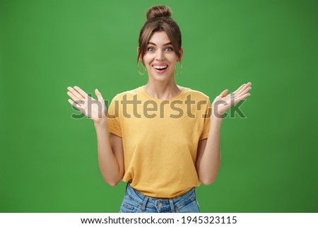 Amazed and surprised woman reacting to astonishing gift raising hands near shoulders smiling happily at camera being delighted and pleased with awesome surprise made by friends over green wall