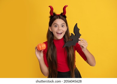 amazed teen girl wearing imp horns holding pumpkin and bat on yellow background, november