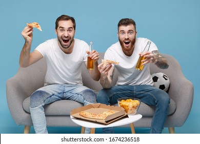 Amazed Two Young Men Guys Friends In White T-shirt Sit On Couch Isolated On Pastel Blue Background. Sport Leisure Concept. Cheer Up Support Favorite Team With Soccer Ball Hold Beer Bottle Eat Pizza