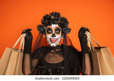 Amazed Woman In Black Halloween Costume And Creepy Makeup Holding Shopping Bags Isolated On Orange