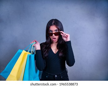 Amazed woman with brown hair, in black jeans and jersey carries blue and yellow shopping bags while she looks above her sunglasses.