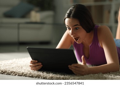 Amazed woman in the night using tablet lying on the floor at home