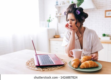 Amazed wondered young woman look on laptop. Watching movie in kitchen. Croissant and cup of drink on table. Careless housekeeper in room. Life withour work. Daylight in morning.