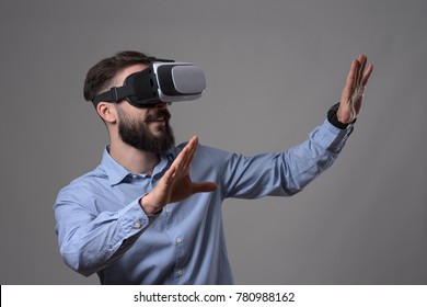 Amazed young adult bearded smart casual man wearing virtual reality vr headset gesturing hands and looking at copyspace over gray background.