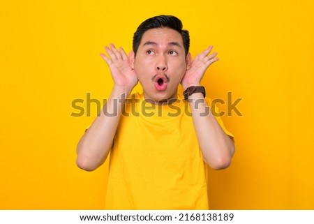 Amazed young Asian man in casual t-shirt holding hands near his ears isolated on yellow background. People lifestyle concept