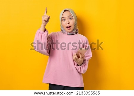 Amazed young Asian woman in pink shirt holding mobile phone and pointing fingers up, having a good idea on yellow background
