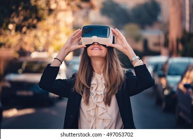 amazed young beautiful girl testing virtual reality 3D video glasses VR headset dressed in a office outfit curious by augmented reality on the street with beautiful autumn sun light colors - Powered by Shutterstock