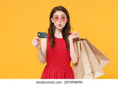 Amazed young brunette woman girl in red summer dress eyeglasses posing isolated on yellow background studio. People lifestyle concept. Hold package bag with purchases after shopping credit bank card