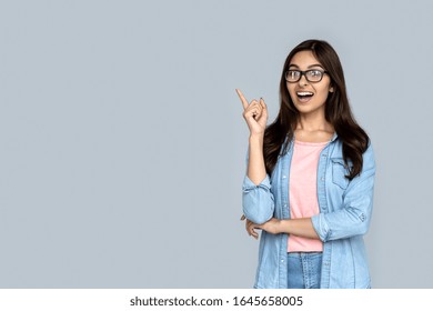 Amazed young happy indian girl student lady shopper professional excited face looking at camera pointing finger at copy space surprised by shopping sale offer isolated on gray studio background.