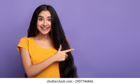 Amazed Young Indian Lady With Long Dark Hair Pointing At Copy Space Over Purple Studio Background. Excited Woman Indicating Free Space, Showing Place For Advert Or Promotional Text