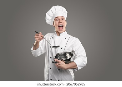 Joven chef asombroso con uniforme y sombrero mirando la cámara con la boca abierta mientras está de pie en estudio gris con salsa y cuchara de sopa