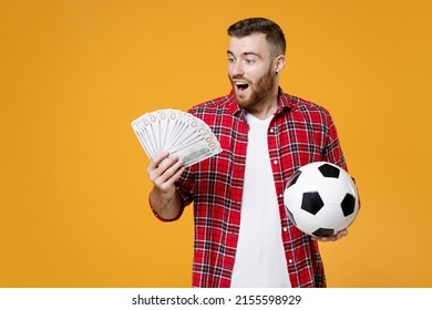 Amazed young man football fan in basic red shirt cheer up support favorite team with soccer ball hold fan of cash money in dollar banknotes isolated on yellow background. People sport leisure concept