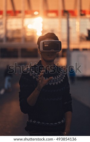 Amazed young man with one hand touching in the air wearing a pair of VR glasses in a cool sweater and trendy outfit impressed by augmented reality sunlit by an amazing sunset reflection fro background