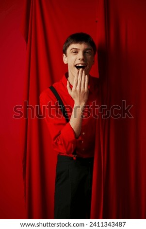amazed young man in suspenders smiling and covering mouth near red vibrant curtain, merriment