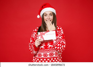 Amazed young Santa woman in sweater Christmas hat hold gift certificate covering mouth with hand isolated on red colour background, studio portrait. Happy New Year celebration merry holiday concept