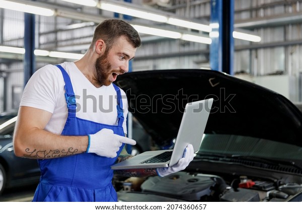 Amazed young technician car mechanic man in\
overalls t-shirt use hold point index finger on laptop pc computer\
make diagnostics fix problem with raised hoodwork in vehicle repair\
shop workshop indoor