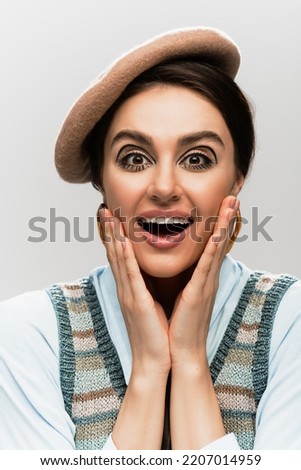 amazed young woman with makeup in beret and knitted vest isolated on grey