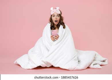Amazed young woman in pajamas home wear sleep mask sit using mobile cell phone keeping mouth open resting at home isolated on pastel pink background studio portrait. Relax good mood lifestyle concept