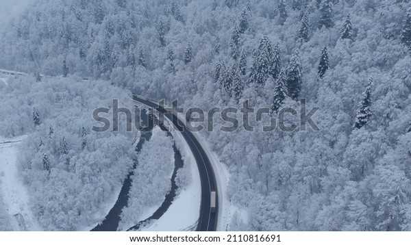 Amazing aerial drone shot: Trucks driving by the\
road in beautiful winter mountains forest completely covered by\
white snow.
