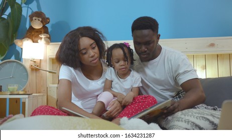 Amazing African American Family Reading Books Educating A Child At Home. Cute Portrait Of Young Parents And Braided Kid Girl Learning Words Lying On Bed.