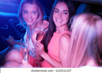 Amazing And Attractive Girls Are Sitting In Car And Looking Outside From Car's Window. Blonde Girl Is Taking Picture On Phone. She And Brunette Girl Are Holding Glasses Of Champagne.