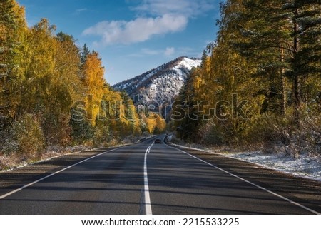 Amazing autumn view with an asphalt road in the mountains, first snow, forest and golden trees on the roadside against a background of blue sky and clouds. Altai, Russia