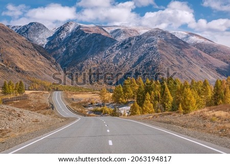 Amazing autumn view with a winding asphalt road, mountains with first snow, forest and golden trees on the roadside against a background of blue sky and clouds. Altai, Russia