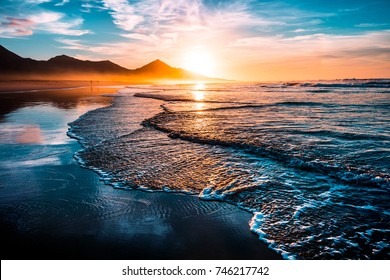 Erstaunlicher Sonnenuntergang am Strand mit endlosem Horizont und einsamen Figuren in der Ferne und unglaublichen schwachen Wellen. Vulkanhügel im Hintergrund.