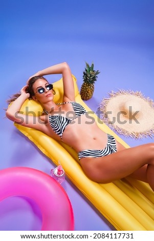  amazing beautiful caucasian woman who wears in bikini lyes on inflatable yellow matress while posing on purple background . Summer concept. Travel concept. Relax concept