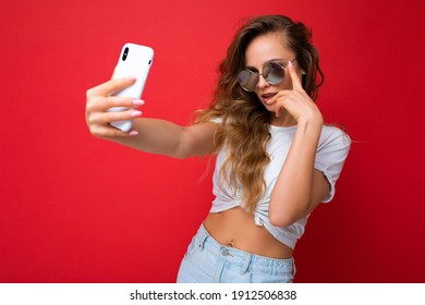 amazing beautiful young blonde woman holding mobile phone taking selfie photo using smartphone camera wearing sunglasses everyday stylish outfit isolated over colorful wall background looking at