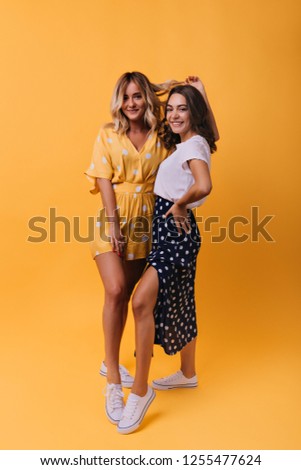 Amazing blonde girl in yellow outfit chilling with best friend. Indoor full-length shot of two enthusiastic sisters smiling to camera.