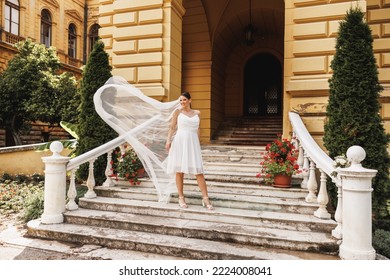 Amazing Bride Posing On The Stairs Outdoor While Her Bridal Long Veil Flying On Wind In Air.