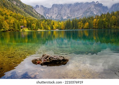 Amazing clear mountain lake in forest among fir trees in sunshine. Bright scenery with beautiful turquoise lake against the background of snow-capped mountains, mountains reflection, Italy