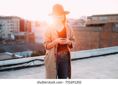 Amazing epic shot of young woman or teenager in coat and fedora hat standing on top of rooftop in big city, uses smartphone to chat or message application during sunset - Powered by Shutterstock