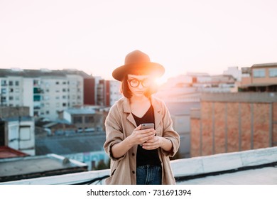 Amazing epic shot of young woman or teenager in coat and fedora hat standing on top of rooftop in big city, uses smartphone to chat or message application during sunset - Shutterstock ID 731691394