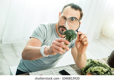 Amazing Facts. Concentrated Handsome Beardful Man Spending Time In The Bright Room Holding A Broccoli Looking Through Magnifying Glass.