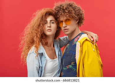 Amazing fashionable girl model with curly ginger hairstyle dressed in jeans shirt posing indoors, hugging young male with crisp hair in t-shirt with flower print, shades and stylish yellow bomber