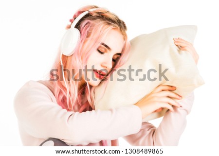 Amazing girl with pink curly hair in headphones playfully posing in cute pajama holding pillow and laughing to camera. Indoor photo of inspired young woman in night-suit isolated on white background