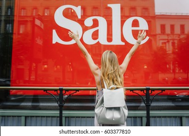 Amazing Girl Raised Hands Next To Shop Window With Sale Ad. Red Sign On A Shop Window. Sale In A Clothing Store