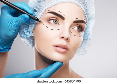 Amazing girl wearing blue medical hat at studio background and looking right, perforation lines on face, close up, plastic surgery concept, doctor's hand in glove making marks on patient's face.