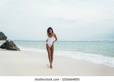 amazing girl in a white swimsuit with a beautiful sports body walking and posing on a white sand beach. A tanned young woman with curly hair is resting and sunbathing. Charming model posing near the