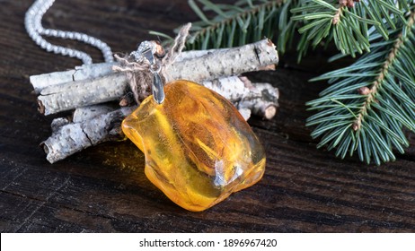 Amazing Glittering Bright Yellow Baltic Amber Pendant Lies On The Birch Branches With Fir Twigs On The Wooden Surface.  Amber Texture,  Ancient Good Luck Charm. 