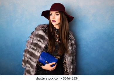 Amazing Gorgeous Lady In Elegant Black Dress, Fluffy Coat And Stylish Maroon Hat Holding Trendy Blue Clutch Bag. Fashionable Model Posing On Isolated Background, Looking Sideways. Elegance And Beauty.