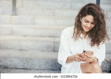 Amazing lovely young woman sitting alone on steps ouside. Look at smartphone in hands and smile. Wear stylish white blouse. Modern technologies and gadgets