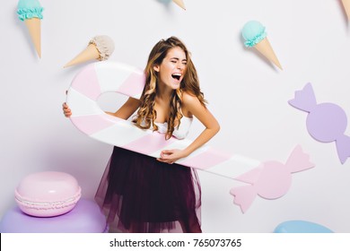 Amazing sweet-tooth girl surrounded by toy sweets having fun on party and singing. Portrait of young woman in elegant violet dress holding big candy cane and posing on decorated background.