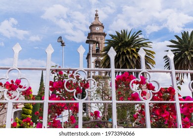 Amazing View From Behind White Metal Fence And Pink Flowers Of Old Bell Tower And Two Palm Trees. Ancient Building And Nature With Beautiful Cloudy Sky As Background. Classic Mexican Architecture