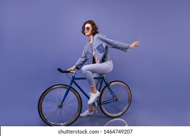 Amazing Woman In Spring Clothes Sitting On Bicycle. Indoor Portrait Of Lovely Girl In Sunglasses Fooling Around On Violet Background.