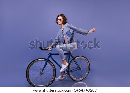 Amazing woman in spring clothes sitting on bicycle. Indoor portrait of lovely girl in sunglasses fooling around on violet background.
