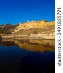 The Amber Fort, located in Amer, Rajasthan, India. The fort is situated on a hill and is reflected beautifully in the calm waters of a lake below. The structure features classic Indian architecture.