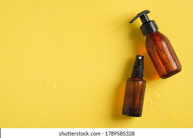 Amber glass cosmetic bottles in droplets of water. Refreshing beauty products on yellow background. Top view, flat lay. 